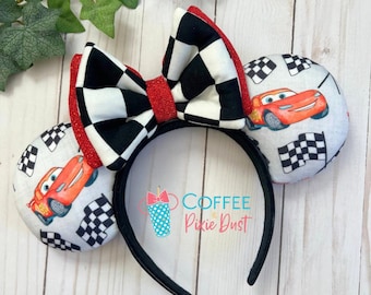 May include: Red race car themed Minnie Mouse ears with a black and white checkered bow. The ears are made of fabric and have a white background with red race cars and checkered flags.