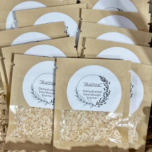 May include: A collection of brown paper pouches with clear windows, each containing a dehydrated sourdough starter. White circular labels with the text "Ruthie" and "Dehydrated Sourdough Starter" are affixed to the pouches. The pouches are arranged in a woven basket.