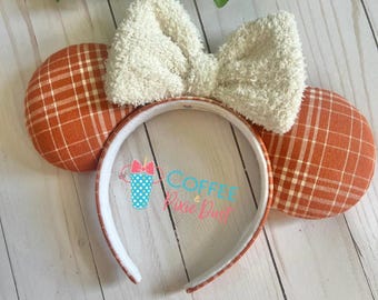 May include: A headband with orange plaid ears and a fluffy cream-colored bow. The headband is white with a matching orange plaid trim. The image includes the text "Coffee Pixie Dust" in a script font.