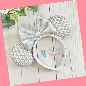 May include: Silver glitter Minnie Mouse ears with a silver bow and a white headband. The ears are decorated with a geometric pattern.