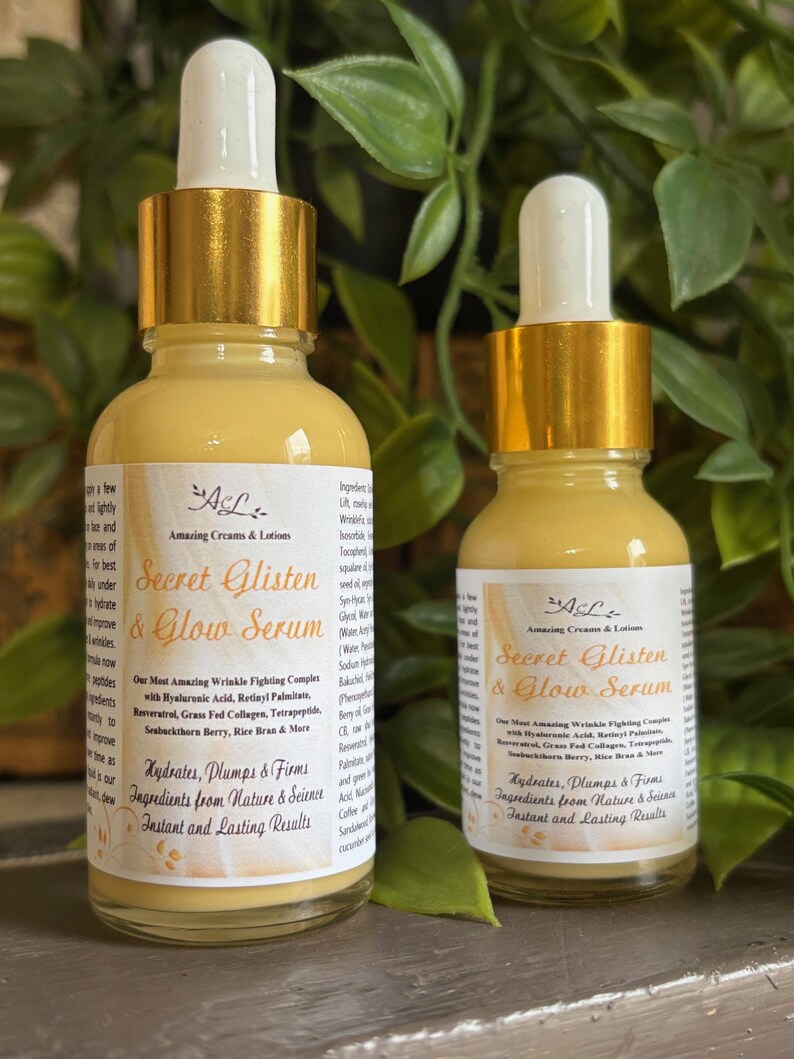 May include: Two amber glass bottles with gold caps and droppers filled with a yellow serum. The bottles are labeled "Secret Glisten & Glow Serum" with the brand name "Amazing Creams & Lotions". The bottles are on a wooden surface.