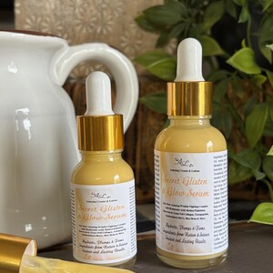 May include: Two amber glass bottles with gold lids and droppers filled with a yellow serum. The bottles are labeled "Secret Glisten & Glow Serum" and list ingredients from nature and science for instant and lasting results.
