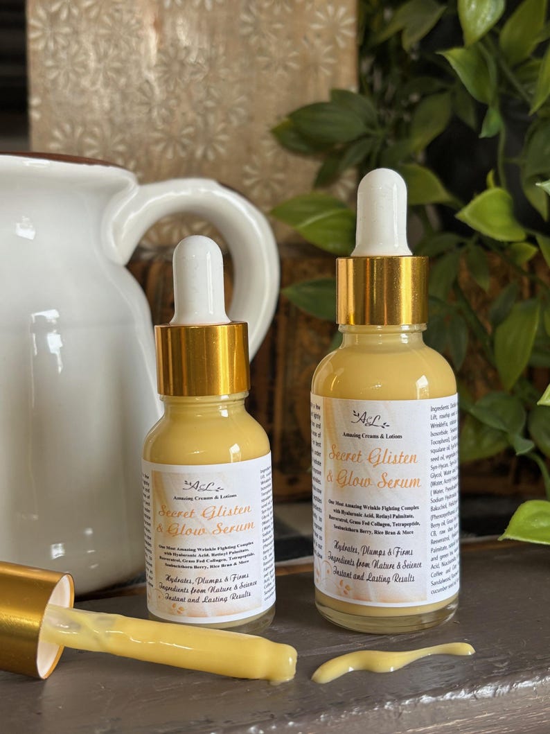 May include: Two amber glass bottles with gold lids and droppers filled with a yellow serum. The bottles are labeled "Secret Glisten & Glow Serum" and list ingredients from nature and science for instant and lasting results.