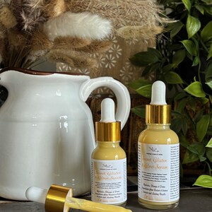 May include: Two amber glass bottles with gold lids and droppers filled with a yellow serum. The bottles are labeled "Secret Glisten & Glow Serum" and contain ingredients that help reduce, plump, and tone.