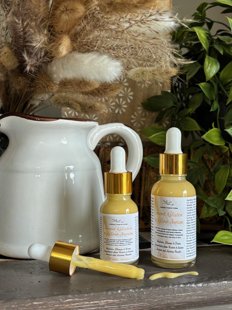 May include: Two amber glass bottles with gold lids and droppers filled with a yellow serum. The bottles are labeled "Secret Glisten & Glow Serum" and contain ingredients that help reduce, plump, and tone.
