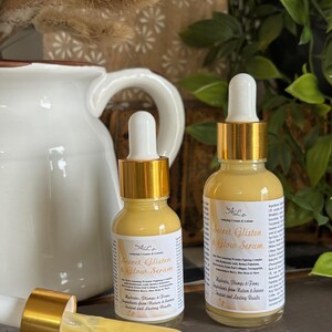 May include: Two amber glass bottles with gold lids and droppers. The bottles are labeled "Secret Glisten & Glow Serum" and contain a serum for skin care. The bottles are sitting on a wooden surface.