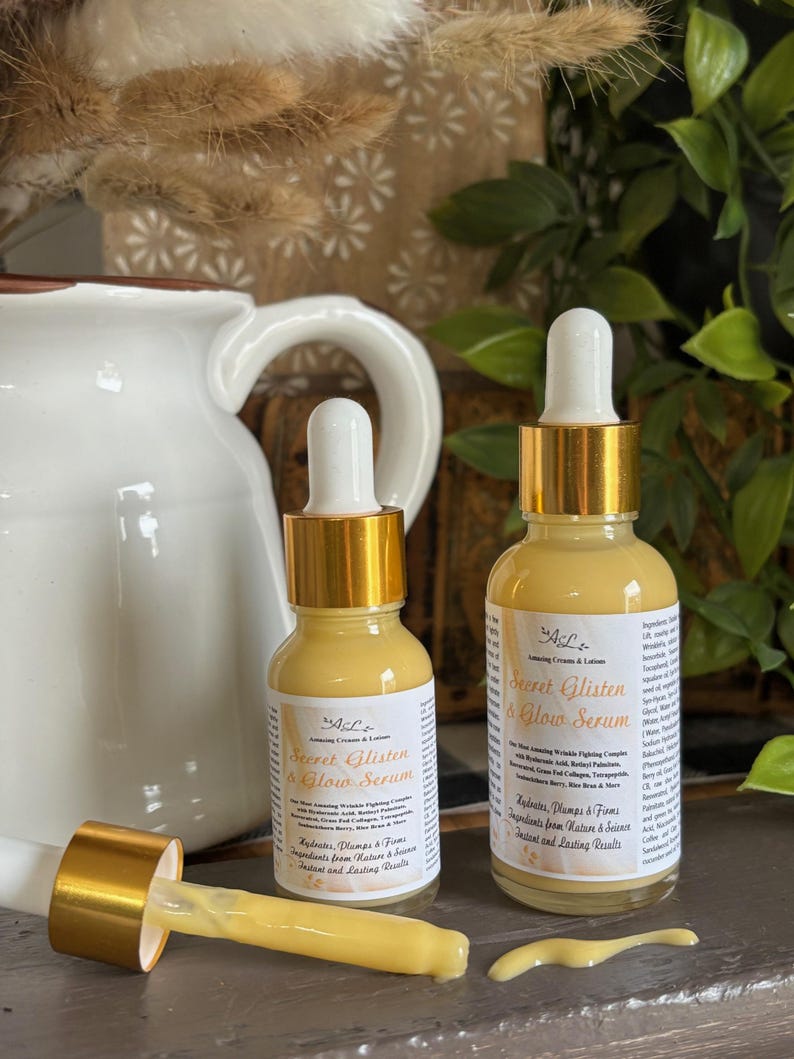 May include: Two amber glass bottles with gold lids and droppers. The bottles are labeled "Secret Glisten & Glow Serum" and contain a serum for skin care. The bottles are sitting on a wooden surface.