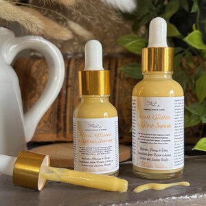 May include: Two amber glass bottles with gold lids and droppers filled with a yellow serum. The bottles are labeled "Secret Glisten & Glow Serum" and contain ingredients from nature for instant and lasting results.