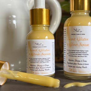 May include: Two amber glass bottles with gold lids and labels that read "Secret Glisten & Glow Serum". The bottles are filled with a yellow liquid. The text on the label says "Hydrates, Plumps & Firms. Ingredients from Nature & Science. Instant and Lasting Results."
