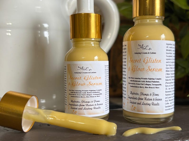 May include: Two amber glass bottles with gold lids and labels that read "Secret Glisten & Glow Serum". The bottles are filled with a yellow liquid. The text on the label says "Hydrates, Plumps & Firms. Ingredients from Nature & Science. Instant and Lasting Results."