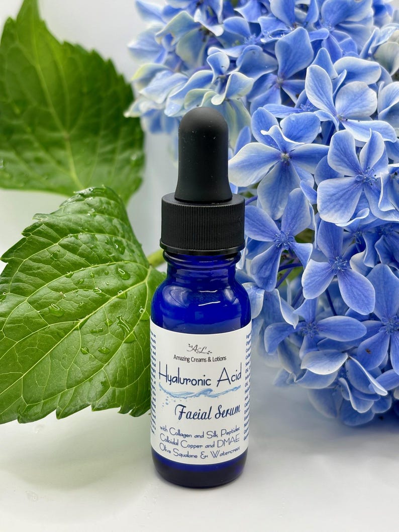 May include: A blue glass bottle with a dropper top, labeled "Hyaluronic Acid Facial Serum" with ingredients listed. The bottle is sitting on a white surface with green leaves and blue flowers in the background.