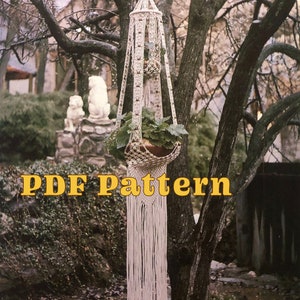 May include: A cream macrame plant hanger, holding a potted plant, hanging from a tree. The design features geometric patterns and beaded details, with long fringe. The text "PDF Pattern" is displayed in yellow.