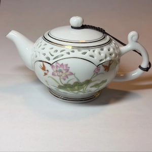 Może przedstawiać: Biały porcelanowy imbryk z kwiatowym i ptasim wzorem. Imbryk ma okrągły korpus z dekoracyjnym ażurowym wzorem wokół góry, zakrzywiony uchwyt i dzióbek. Projekt obejmuje różowe kwiaty lotosu i małe ptaki. Pokrywa jest zabezpieczona czarnym sznurkiem.