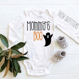 以下が含まれることがあります： 白地に黒とオレンジ色の「Mommy's Boo」の文字が書かれた、長袖のベビーロンパース。文字の右には、白い顔をした黒い幽霊が描かれている。ロンパースは白い木製の表面に置かれ、左には緑の葉の植物がある。