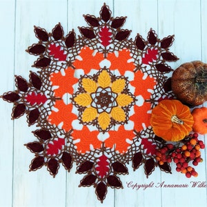 May include: A crocheted doily with a brown, orange, and red leaf design. The doily is surrounded by fall-themed decorations, including pumpkins and berries.