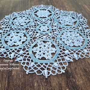 May include: A light blue crocheted doily with a snowflake design. The doily features a central snowflake pattern surrounded by six hexagonal motifs, all connected by a delicate, openwork design. The doily is handmade and has a scalloped edge.