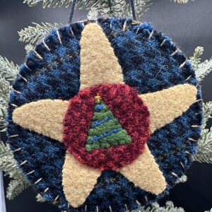 May include: A round, felted wool ornament featuring a dark blue and black plaid background with a large, tan, five-pointed star centered on the front. A textured, deep red circle sits in the middle of the star, displaying a small green Christmas tree.