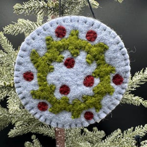 May include: A circular felt ornament in light gray featuring a stitched edge. The center displays a green felt wreath design accented with small, circular dark red felt pieces scattered across the surface. A black string is attached for hanging.