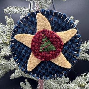 Op de afbeelding: Een rond, donkerblauw, getextureerd wollen ornament met een grote, beige, vijfpuntige ster in het midden. Een kleinere, ronde, donkerrode wollen patch ligt bovenop de ster en toont een groen, driehoekig kerstboomontwerp.