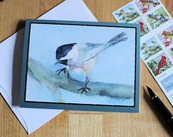 May include: A watercolor painting of a black-capped chickadee perched on a branch. The bird is painted in shades of blue, white, and black, with a light blue background.