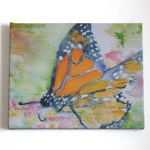 Monarch butterfly watercolor painting printed on canvas that has been stretched taut on wooden frame.  Horizontally formatted, the butterfly fills the frame in shades of orange, deep blue, black and white.  The insect rests on loosely painted flowers