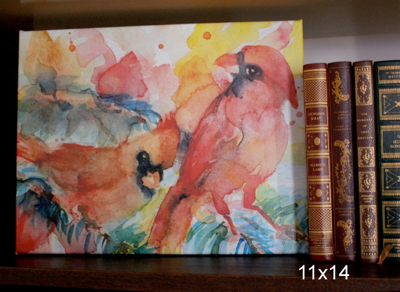 Stretched canvas print of colorful cardinals is displayed on a book shelf for size reference. The print is a few inches taller than a standard size hardcover book.  Text reads 11x14.