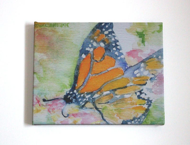 Monarch butterfly watercolor painting printed on canvas that has been stretched taut on wooden frame.  Horizontally formatted, the butterfly fills the frame in shades of orange, deep blue, black and white.  The insect rests on loosely painted flowers