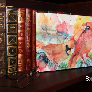 Stretched canvas print of colorful cardinals is displayed on a book shelf with a pair of eyeglasses for size reference. The print is shorter than a standard size hardcover book.  Text reads 8x10.