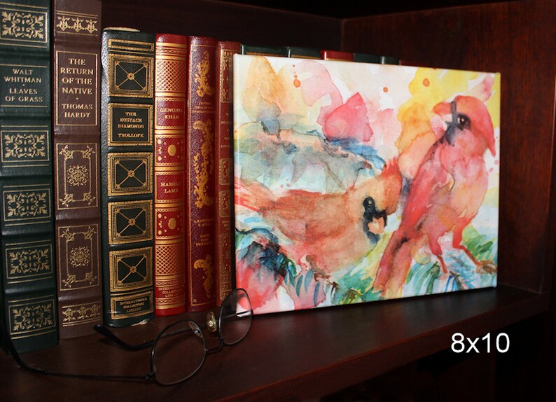 Stretched canvas print of colorful cardinals is displayed on a book shelf with a pair of eyeglasses for size reference. The print is shorter than a standard size hardcover book.  Text reads 8x10.