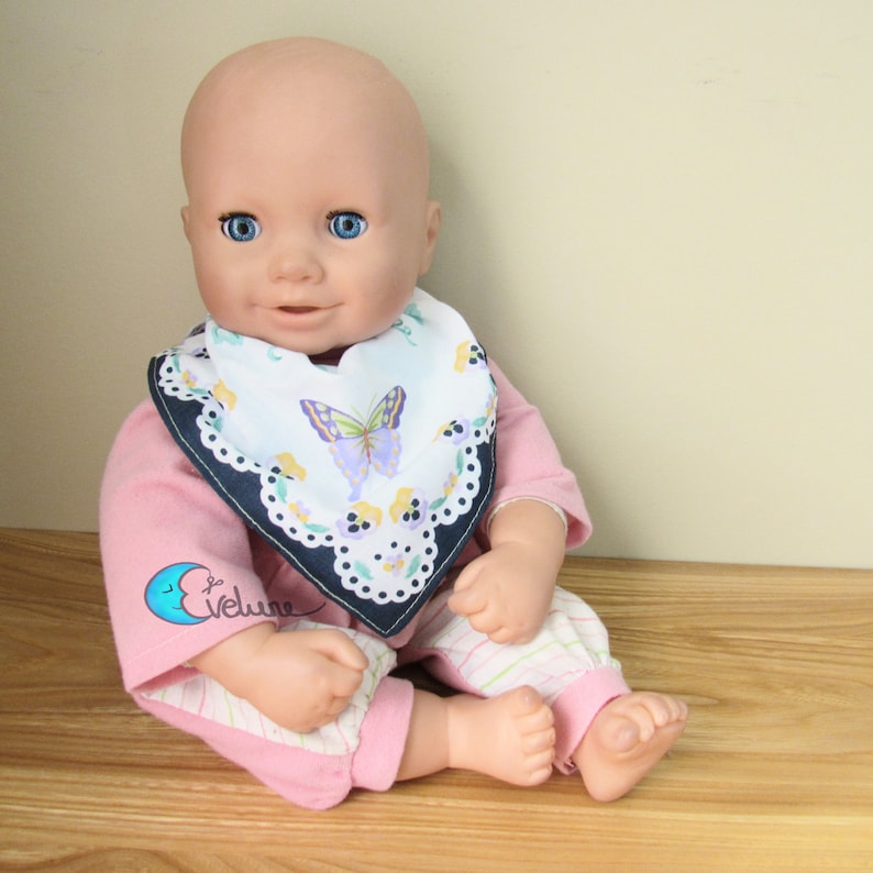 May include: A baby doll with blue eyes, wearing a pink long-sleeved shirt and striped pants. The doll has a white bib with a butterfly design and a navy blue border. The doll is sitting on a wooden surface.