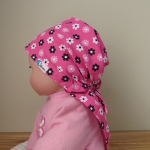 K&ouml;nnte beinhalten: Ein pinkfarbenes Kopftuch mit einem Blumenmuster aus wei&szlig;en und schwarzen Bl&uuml;ten. Das Kopftuch ist am R&uuml;cken gebunden und befindet sich auf einer Puppe. Die Puppe tr&auml;gt ein pinkfarbenes Oberteil.