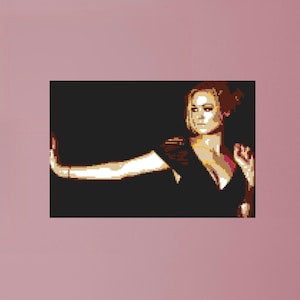 May include: A woman with long brown hair is wearing a black dress and posing with her arm extended. The background is black.