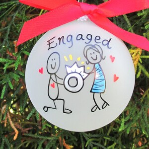 Pu&ograve; includere: Una palla di Natale di vetro trasparente con la scritta "Engaged" e un'illustrazione disegnata a mano di una scena di proposta. L'ornamento ha un nastro rosso ed &egrave; appeso a un gancio dorato.