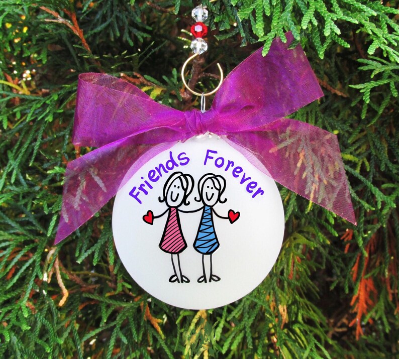 May include: A white ornament with the words "Friends Forever" and a drawing of two stick figure friends. The ornament is decorated with a purple ribbon and hangs from a gold hook with red and clear beads. The background is green.