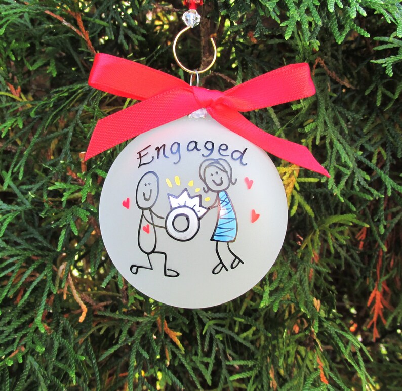 Pu&ograve; includere: Una palla di Natale di vetro trasparente con la scritta "Engaged" e un'illustrazione disegnata a mano di una scena di proposta. L'ornamento ha un nastro rosso ed &egrave; appeso a un gancio dorato.