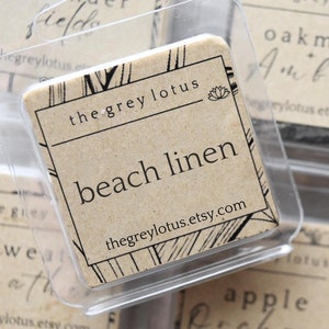 Puede incluir: Primer plano de una colección de pequeñas fundidas de cera cuadradas en recipientes de plástico transparente. Cada recipiente tiene una etiqueta con un nombre de aroma diferente y el texto "thegreylotus.etsy.com". Los aromas incluyen "beach linen", "oakmoss amber", "apple orchard", y "saffron cedar".