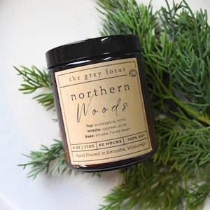 May include: A brown glass candle jar with a black lid and a brown paper label. The label reads "the grey lotus" and "northern Woods". The candle is 8 oz and has a burn time of 60 hours. The candle is made with 100% soy wax and is hand poured in Kenosha, Wisconsin.