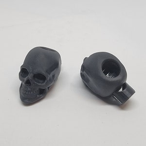 Puede incluir: Dos cierres de palanca de plástico negro en forma de calavera. Un cierre es un diseño de calavera simple, mientras que el otro tiene un agujero en la parte superior y una pequeña pieza de plástico negro unida al lado.