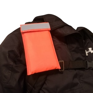 May include: A bright orange safety pouch with a reflective strip attached to a black rucksack.
