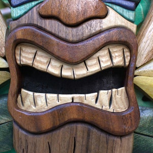 May include: A wooden tiki mask with a wide, toothy grin. The mask is carved with intricate details and painted in shades of brown, green, and yellow.