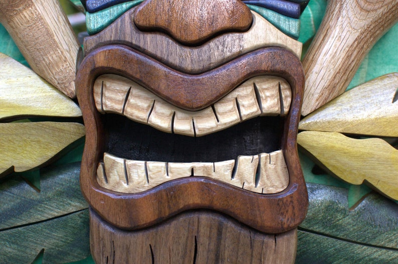 May include: A wooden tiki mask with a wide, toothy grin. The mask is carved with intricate details and painted in shades of brown, green, and yellow.