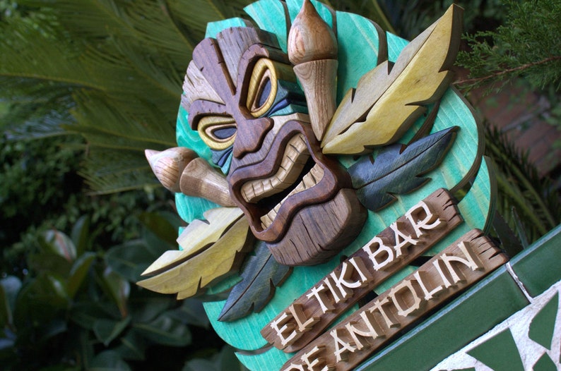 May include: A wooden tiki mask with a carved mouth and eyes, surrounded by carved leaves. The sign reads "El Tiki Bar" and "De Antollin".