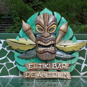 May include: A wooden tiki mask with a carved face and a green, yellow, and brown leaf design. The sign reads "El Tiki Bar de Antolin".