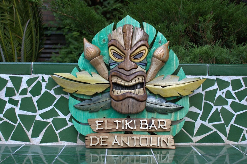 May include: A wooden tiki mask with a carved face and a green, yellow, and brown leaf design. The sign reads "El Tiki Bar de Antolin".