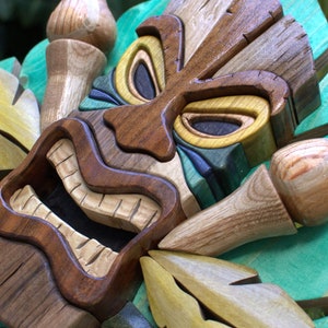 May include: A wooden tiki mask with a carved face and green, yellow, and brown accents. The mask is surrounded by carved wooden leaves.