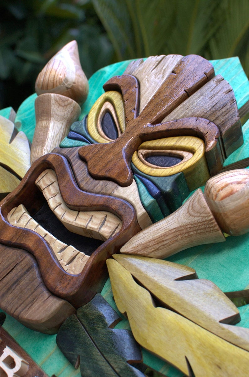May include: A wooden tiki mask with a carved face and green, yellow, and brown accents. The mask is surrounded by carved wooden leaves.