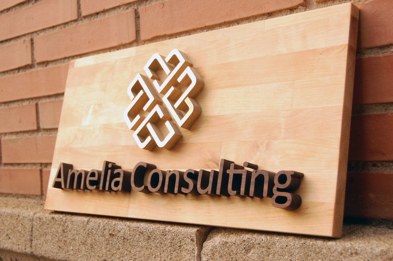 May include: A wooden sign with the text "Amelia Consulting" in raised brown letters. A white and gold geometric design is above the text.