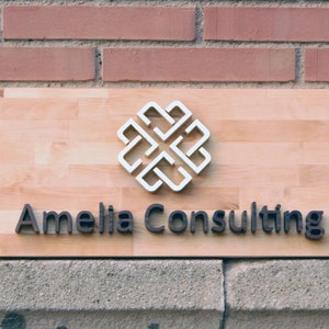 May include: A wooden sign with the text "Amelia Consulting" in black raised letters. A white, stylized, geometric design is above the text.