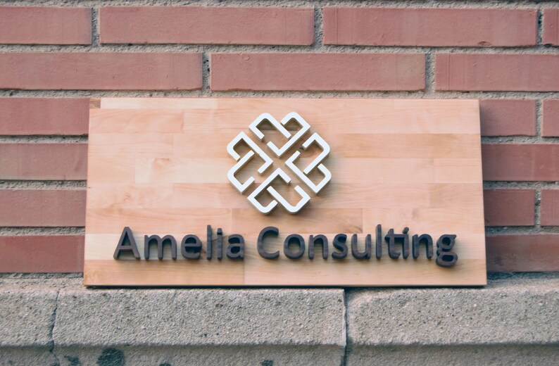 May include: A wooden sign with the text "Amelia Consulting" in black raised letters. A white, stylized, geometric design is above the text.