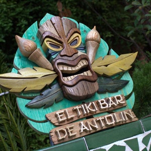 May include: A wooden tiki mask with carved details and a green background. The sign reads "El Tiki Bar de Antolin".
